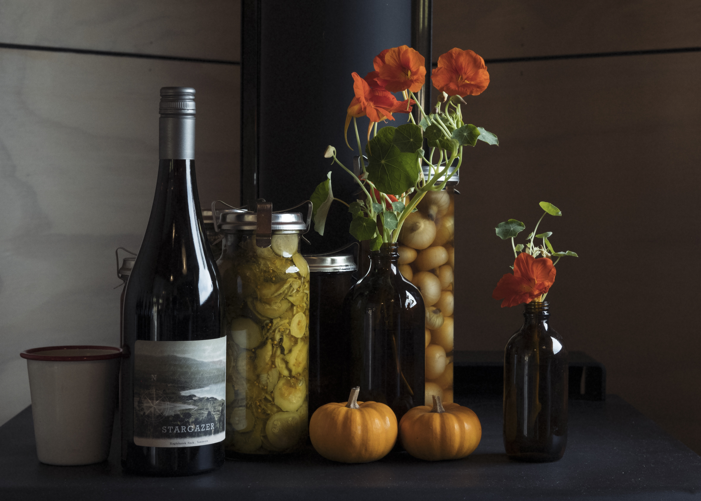 Wine bottle on table with small pumpkins, bottle of pickled vegetables,  and bottles with flowers